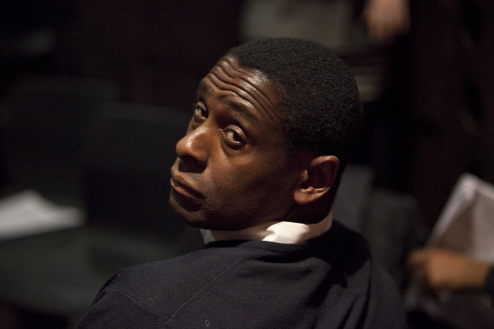 Actor David Harewood sits in a white shirt and black jumper. His face is turned to face the camera with eyebrows raised.