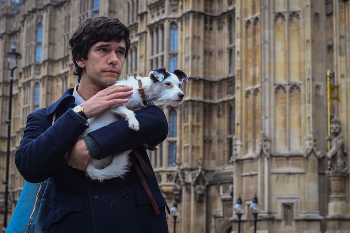 A scene from BBC's A Very English Scandal. Ben Whishaw, looking pensive, stands in front of the Houses of Parliament holding a scruffy white terrier.