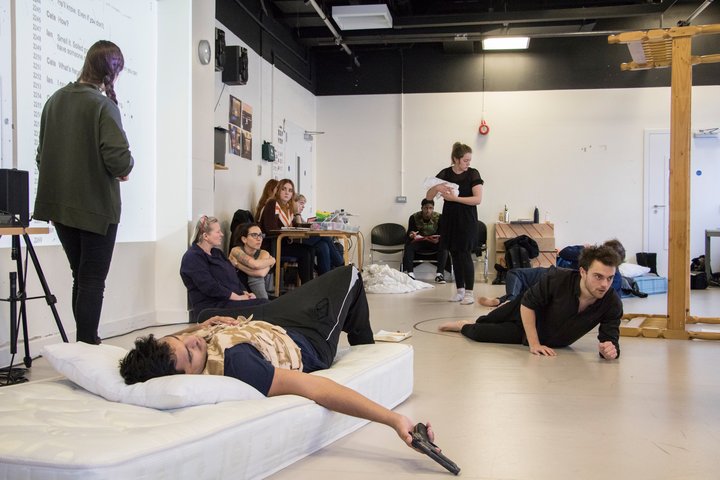 A group of people including actors, directors are working on a scene. There is a man lying on  a bed in the middle lazily holding a gun.