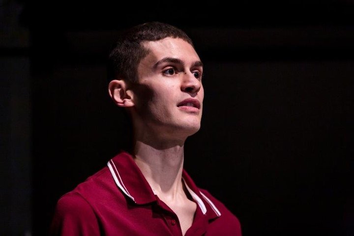 A head and shoulders view of actor Josh Zare wearing a dark red polo shirt in front of a black backdrop, on stage at the Orange Tree Theatre