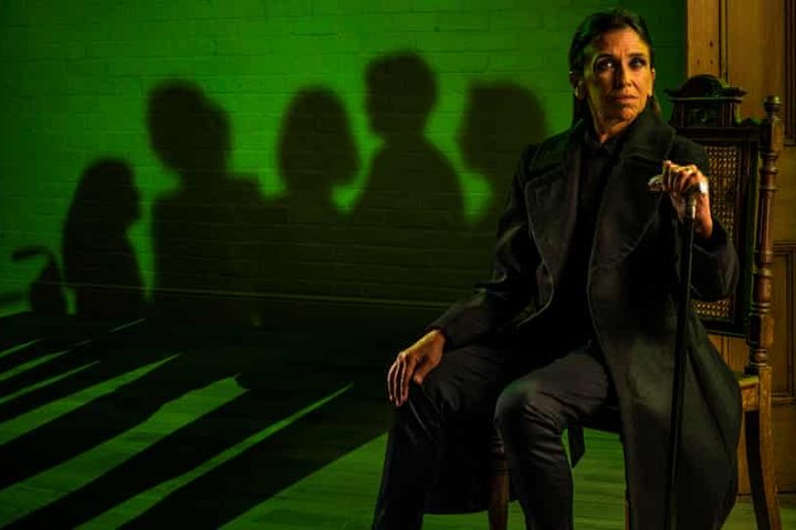 Kathryn Hunter sits on stage wearing black against an acid green brick wall in a scene from The House of Bernarda Alba