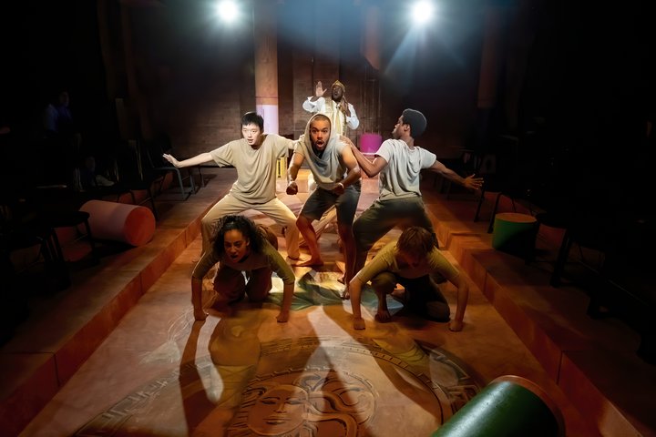 Eight students perform on stage in dramatic lighting. The image is energetic with students braced in a chariot formation.