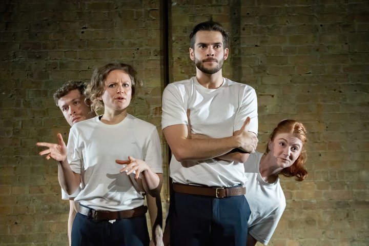Four actors on stage in white t-shirts with inquisitive and serious expressions.