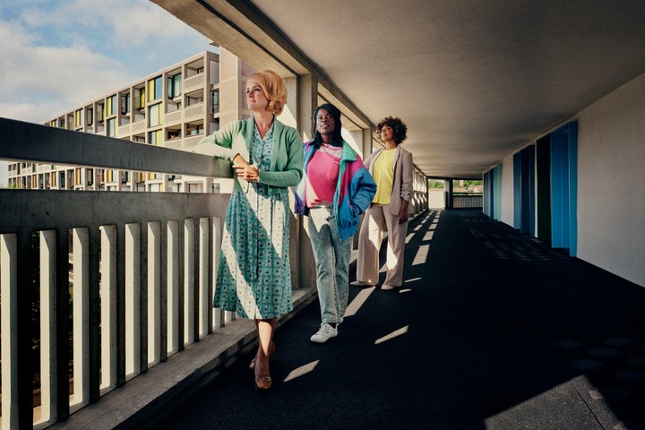 Three actors standing in walkway of tower block with striking lighting.