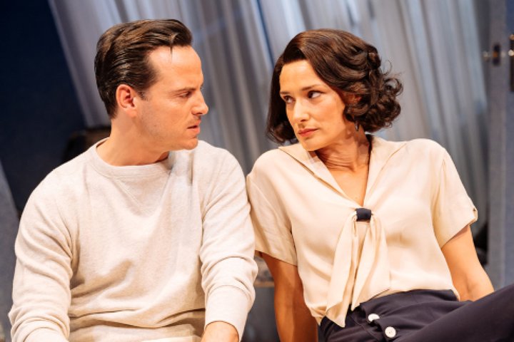 Andrew Scott and Indira Varma sit clsoe together on a couch looking into each other's eyes sceptically in a performance of Present Laughter