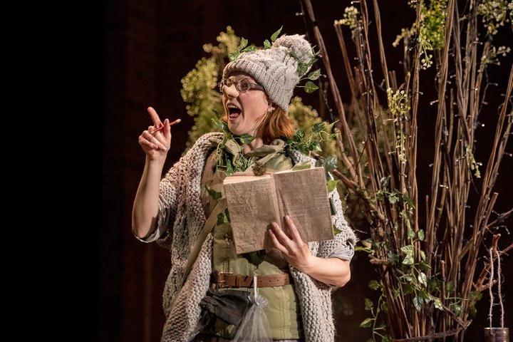 A women dressed like an elf pointing to a plant in a forest on stage