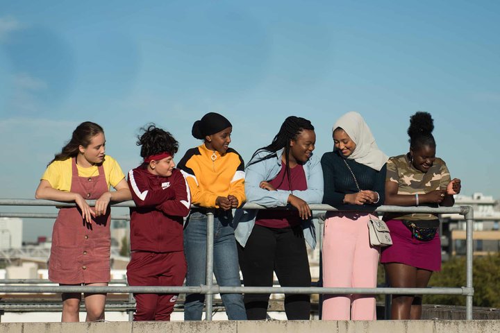 In a scene from the film Rocks, six girls stand in a line leaning against a railing in a city setting against a bright blue sky, smiling together.
