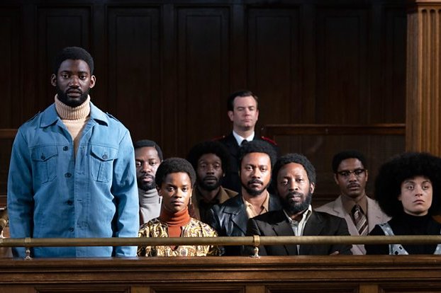 In a scene from the film Mangrove, a group of black men and women including RADA grads Shaun Parkes sit in the dock a dark wooden court room.