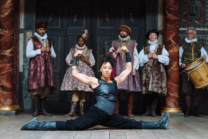 Actress Stacy Abalogun on stage at the Globe playing a wrestler. She is in the splits with her arms raised above her head, looking strong and powerful