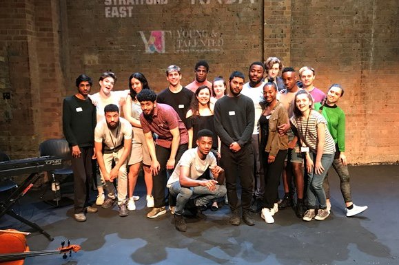 A group of young people in the late teens and early 20s stand smiling, grouped in front of a brick wall at the back of RADA's theatre,