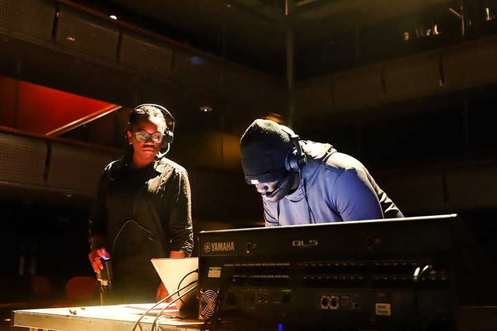 A young technician in a dark beanie hat works at a sound desk. Another young technician looks over the work with a headset on.